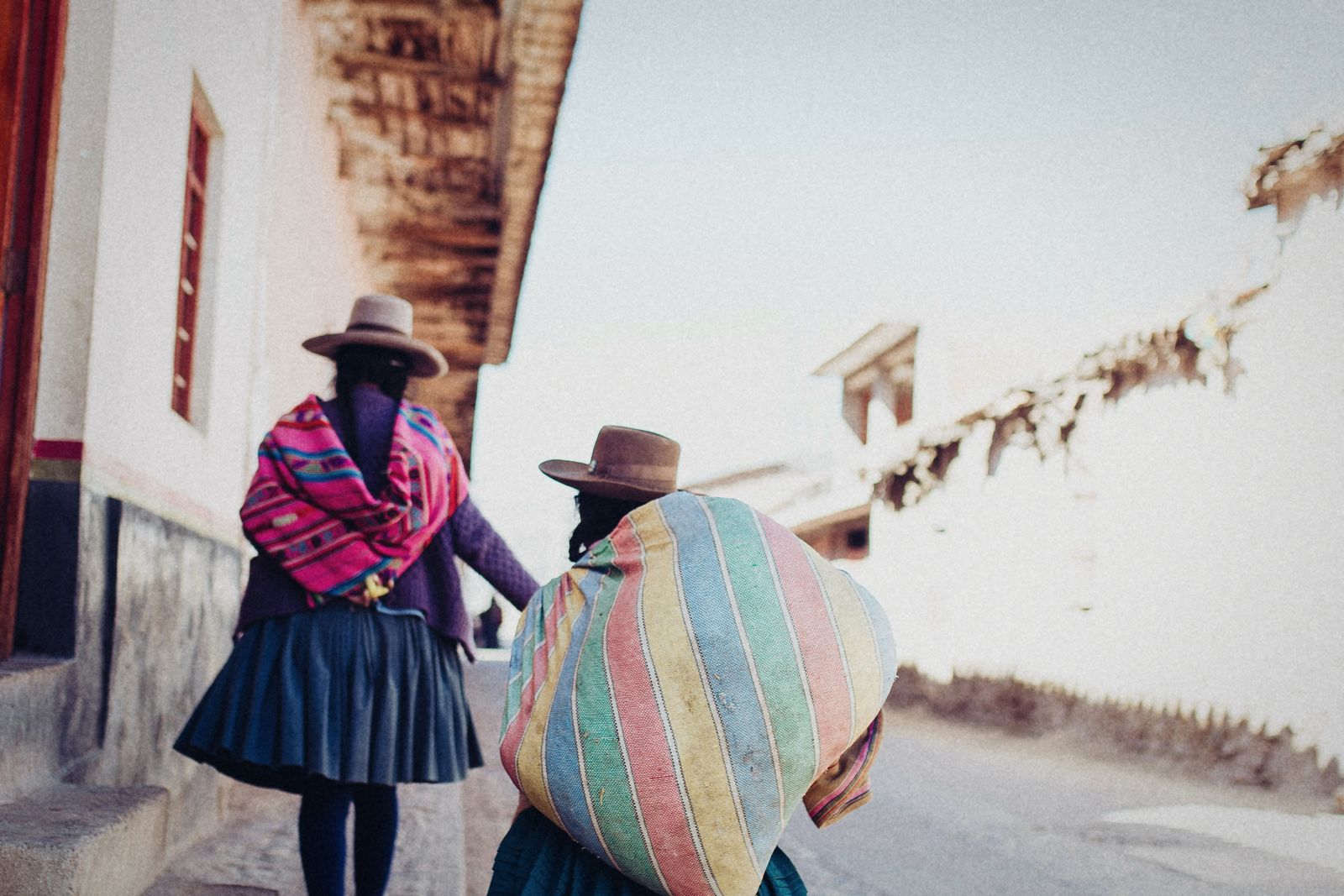 Dos mujeres andinas, de espaldas, suben por una calle empedrada de Cusco cargando bultos en la espalda